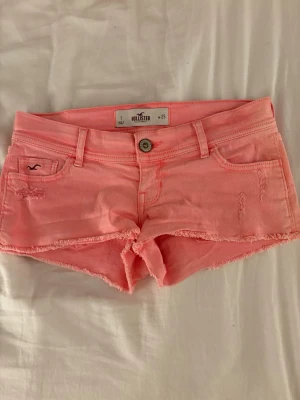 Rosa jeansshorts från Hollister - Säljer ett par ljusrosa jeansshorts från Hollister med låg midja och slitna detaljer. Shortsen har fem fickor, bälteshällor och knappgylf. Perfekta för varma dagar och ger en avslappnad vibe.