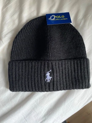 Svart ribbstickad mössa Polo Ralph Lauren - Snygg svart ribbstickad mössa från Polo Ralph Lauren med klassisk broderad logga framtill. Mössan har uppvikt kant och är tillverkad i mjukt material som håller dig varm. Perfekt accessoar för kalla dagar och enkel att matcha med olika outfits.
