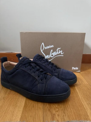 Mörkblå sneakers från Christian Louboutin stl40 - Snygga mörkblå sneakers från Christian Louboutin i mocka med klassisk röd sula. Lågt skaft, svarta skosnören och diskret design som ger en lyxig känsla. Perfekta för dig som vill ha något exklusivt och stilrent ej låda till