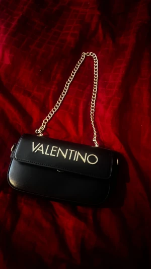 Svart axelväska från Valentino - PRIS GÅR ATT DISKUTERA!  Snygg svart axelväska från Valentino med guldigt kedjehandtag och stort logotryck på framsidan. Väskan har en stilren, rektangulär form och är tillverkad i slätt skinn. Perfekt för dig som gillar lyxiga detaljer och vill sticka ut med en ikonisk accessoar.