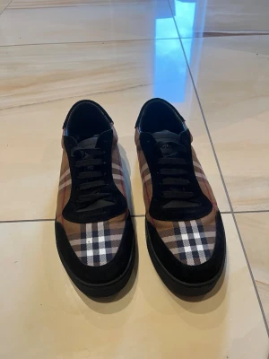 Burberry rutiga sneakers låga - Snygga låga sneakers från Burberry med klassiskt beige, svart, vitt och rött rutmönster. Skorna har svarta mockadetaljer och svarta snören samt en svart sula. Perfekta för dig som vill ha en stilren och tidlös look med lyxig känsla.