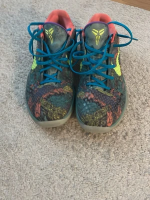 Nike Kobe färgglada basketskor - Säljer ett par Nike Kobe 6 Protro sneakers med färgstarkt mönster i grönt, orange och blått. Skorna har blå skosnören, neongult Swoosh och Kobe-logga på plösen. Låg modell med syntetmaterial och textilfoder. Perfekta för dig som vill sticka ut på basketplanen. Jag har burit dem men de är i bra skick.