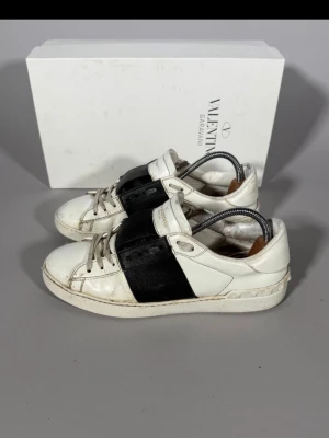 Valentino vita sneakers med svart detalj - Snygga vita sneakers från Valentino Garavani med bred svart läderdetalj över mitten. Klassisk låg modell med vita snören och diskret logga på plösen. Tillverkade i läder för en exklusiv känsla och stilren look.