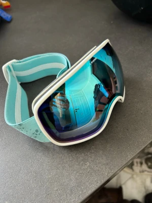 Blå Everest skidglasögon - Snygga skidglasögon från Everest med blått, spegelreflekterande glas och vitt ram. Justerbart turkost band med logga och vita detaljer. Perfekta för skidåkning eller snowboard och ger bra skydd mot snö och vind. Passar 10-15 år