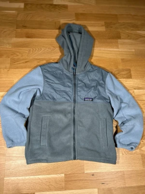 Blå fleecehoodie från Patagonia - Mysig blå hoodie från Patagonia med dragkedja och två fickor framtill. Tillverkad i mjuk fleece med förstärkt bröstparti och huva. Perfekt för chill dagar eller när du vill ha något bekvämt och stilrent. Klassisk Patagonia-logga på bröstet.