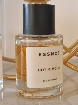ESSNCE Hot Mimosa Eau de Parfum - En stilren parfymflaska från ESSNCE med doften Hot Mimosa. Flaskan är cylindrisk i genomskinligt glas med en svart matt kork och en enkel vit etikett med svart text. Innehållet har en varm, bärnstensfärgad ton. Perfekt för dig som gillar moderna och fräscha dofter.