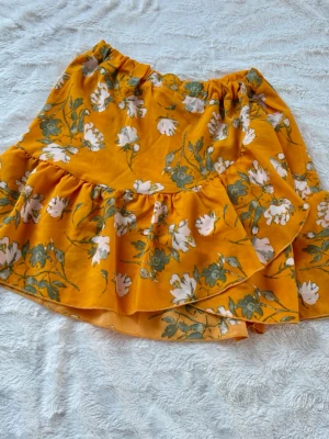 Gul blommig volangkjol - Superfin kort kjol i starkt gul färg med blommigt mönster och volangdetalj. Kjolen har resår i midjan och är perfekt för dig som gillar färg och mönster. Materialet känns lätt och luftigt, perfekt för varmare dagar.