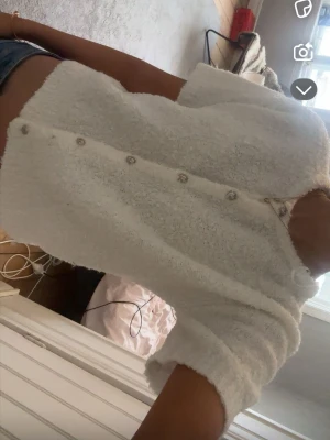 Vit fluffig kofta med knappar - Mysig vit kofta i fluffigt material med dekorativa silverfärgade knappar längs framsidan. Koftan har en avslappnad passform och långa ärmar, perfekt för lager på lager. Enkel och stilren design som passar till det mesta.