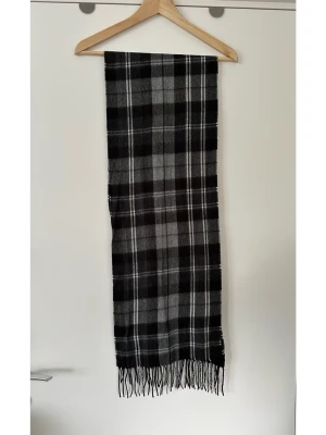 Grå svart rutig halsduk från DAKO - Snygg halsduk från DAKO i svart och grått rutigt mönster med tunna vita linjer. Halsduken har klassiska fransar i ändarna och är tillverkad i ett mjukt tyg som känns skönt mot huden. Perfekt accessoar för att lyfta din outfit med en tidlös look. Mycket gott skick, som ny. Nypris 699:-