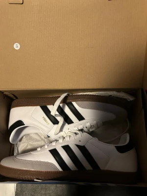 Adidas Samba OG vita och svarta sneakers - Klassiska Adidas Samba OG sneakers i vitt läder med svarta ränder och brun gummisula. Snygg retrostil med gulddetaljen 'SAMBA' på sidan. Perfekta för dig som gillar streetwear och tidlös design. Skorna har låg profil och ikoniska detaljer.