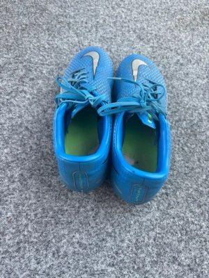 Blå Nike fotbollsskor med snörning - Säljer ett par blå fotbollsskor från Nike med vita detaljer och stora Nike-loggor på ovansidan. Skorna har snörning och en låg profil, samt en grön insida. Perfekta för dig som vill ha snygga och funktionella skor på planen.