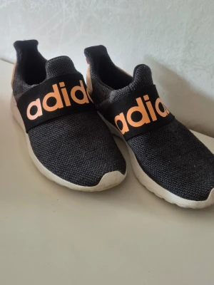 Svarta Adidas slip-on sneakers - Snygga svarta slip-on sneakers från Adidas med stor logga i orange på bred resår över foten. Ovandelen är i mesh-tyg för bra andningsförmåga och sulan är vit och mjuk för skön komfort. Perfekta för en avslappnad och sportig stil.Stl:37 Är i jätte bra skick använda endast ett fåtal tal ggr.Super sköna att ha på.