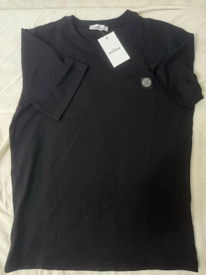 Svart t-shirt från Stone Island - Snygg svart t-shirt från Stone Island med klassisk logopatch på bröstet. T-shirten har rund hals och korta ärmar, perfekt för en clean och stilren look. Tillverkad i mjuk bomull för skön komfort.
