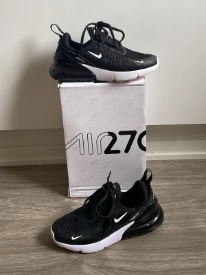 Nike Air Max 270 svarta sneakers - Snygga svarta Nike Air Max 270 sneakers med vit sula och tydlig Air-enhet i hälen. Ovandelen är i mesh för bra ventilation och har vita detaljer som swoosh-loggan och texten 'AIR270' på sidan. Skorna har snörning och en sportig, modern look. ÄKTA!