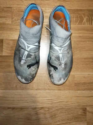 Puma match 7 svart Puma logga - Säljer ett par gråa fotbollsskor från Puma med svart logga på ovansidan och stora blå detaljer på sidorna. Skorna har orange innersula och rosa dobbar. Ovandelen är i textilmaterial och designen är modern med snörning över foten.