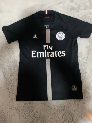 PSG Neymar Jr matchtröja svart (ÄKTA)  - Säljer en svart PSG matchtröja med Neymar Jr och nummer 10 på ryggen. Tröjan har Fly Emirates-tryck på bröstet, PSG-logga och Jordan-logga framtill samt en vit rand i mitten. Perfekt för dig som älskar fotboll och vill stötta Paris Saint-Germain.