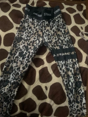 Leopardmönstrade leggings från Stronger - Snygga leggings med leopardmönster från Stronger. Hög midja med logga runt linningen och svart detalj med text på ena benet. Stretchigt material som sitter skönt och formar sig efter kroppen. Perfekta för dig som vill sticka ut på gymmet eller i vardagen.
