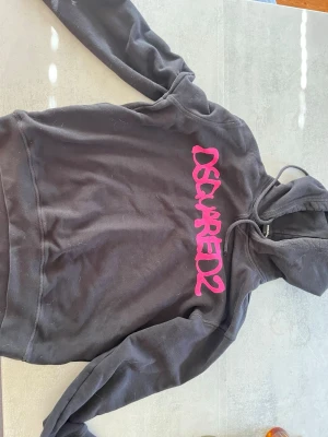 Svart hoodie från Dsquared2 - Säljer en svart hoodie från Dsquared2 med stor rosa logga framtill. Hoodien har huva med dragsko och långa ärmar. Materialet känns mjukt och skönt, perfekt för en chill och avslappnad stil. Passar dig som gillar streetwear och vill sticka ut med färgglad logga.