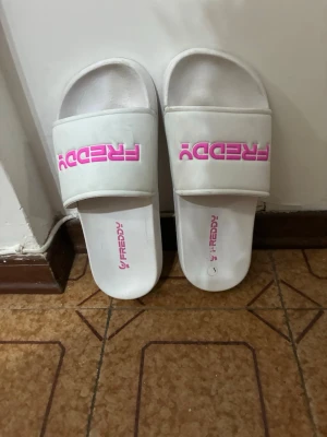 Vita tofflor med rosa FREDDY-logga - Säljer ett par vita tofflor från FREDDY med bred rem och tydlig rosa logga på ovansidan. Tofflorna har en klassisk slip-on design och är tillverkade i syntetmaterial, perfekta för att chilla hemma eller på gymmet.