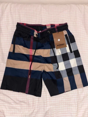 Rutig Burberry shorts med snörning - Säljer ett par snygga Burberry shorts med klassiskt rutigt mönster i svart, beige, marinblått och vitt. Shortsen har elastisk midja med snörning för skön passform och är perfekta för sommaren. Ikonisk design som sticker ut.