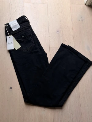 Svarta bootcut jeans från Miss Me - Snygga svarta jeans från Miss Me med bootcut-modell och mid-rise midja. Jeansen har klassiska fickor och dekorativa sömmar. Perfekta för dig som gillar en stilren look med lite edge.                                                                    Midjebredd: 39cm Längd: 111cm