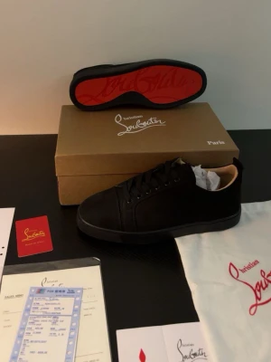 Loubs - Stilrena svarta sneakers från Christian Louboutin med klassisk röd sula och diskret design. Skorna har lågt skaft, svarta skosnören och ovandel i mocka. Perfekta för dig som vill ha en lyxig och clean look.