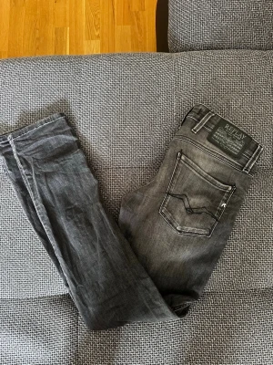 Grå anbass jeans från Replay - Snygga grå slim fit jeans från Replay med klassisk femficksdesign och svarta detaljer. Jeansen har en tvättad look och tydliga sömmar på bakfickorna. Perfekta för dig som gillar en stilren och modern vibe.