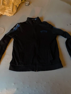 Svart ziptröja från BOSS - Svart ziptröja från BOSS med blå logga på bröstet och blå detaljer längs ärmarna. Tröjan har hög krage, hel dragkedja och två fickor framtill. Perfekt för en sportig och stilren look.