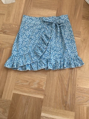 Blå blommig omlottkjol med volang - Supersöt blå kortkjol med vitt blommönster. Kjolen har omlottdesign med volangkant och en knytning i sidan. 