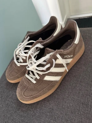 Adidas Spezial - Bruna orginal snöre medföljer (oanvända)       Säljer ett par bruna Adidas Spezial sneakers i mocka med vita detaljer och klassiska tre ränder på sidan. Skorna har vita skosnören och en gummisula som ger retrovibbar. Perfekta för dig som gillar streetwear och sportig stil.
