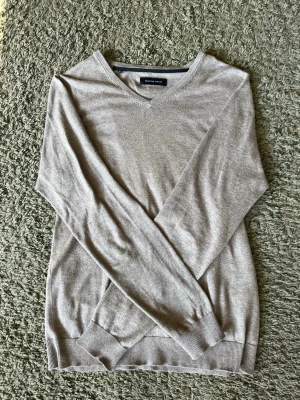 Beige V-ringad tröja - Snygg beige V-ringad tröja med lång ärm och ribbade muddar. Perfekt att bära över en t-shirt för en clean och enkel look. Materialet känns mjukt och lätt, vilket gör den skön att ha på sig hela dagen.
