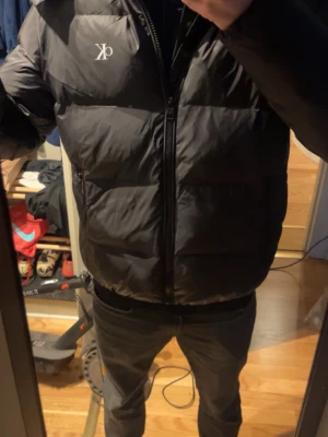 Svart pufferjacka från Calvin Klein - Snygg svart pufferjacka från Calvin Klein med diskret CK-logga på bröstet. Jackan har dragkedja framtill, två sidofickor och hög krage. Tillverkad i ett glansigt material som ger en modern look och håller dig varm under kalla dagar.