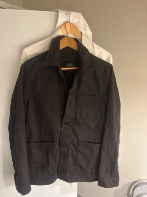 Mörkbrun overshirt med fickor - Stilren mörkbrun overshirt med tre framfickor och knäppning framtill. Klassisk krage och raka ärmar med knapp vid ärmslut. Perfekt lager-på-lager-plagg i ett robust bomullstyg som ger en avslappnad men snygg look.
