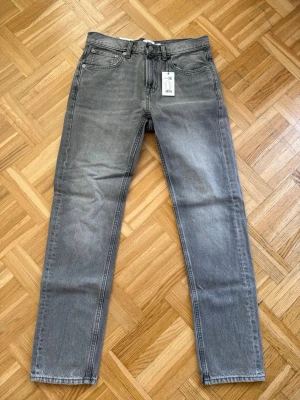 Grå raka jeans New Classic 30/32 - Säljer ett par grå jeans i modellen New Classic med raka ben och klassisk femficksdesign. Jeansen har normal midja och är tillverkade i ett slitstarkt denimtyg. Perfekta för dig som gillar en enkel och stilren look. Helt nya har aldrig använts. 