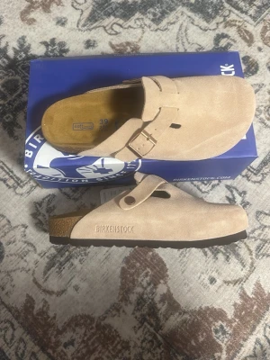 Beige Birkenstock tofflor  - Säljer ett par snygga beige tofflor från Birkenstock i mjuk mocka. De har en klassisk slip-in modell med justerbart spänne och korksula för extra komfort. Perfekta för dig som vill ha både stil och bekvämlighet i vardagen.