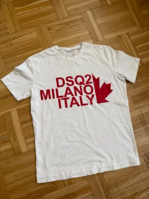 Vit DSQ2 Milano Italy t-shirt - Snygg vit t-shirt med röd text 'DSQ2 MILANO ITALY' och ett rött lönnlöv på bröstet. Klassisk rund hals och korta ärmar. Perfekt för dig som gillar stilrena plagg med statement-tryck. Storlek S. Helt ny