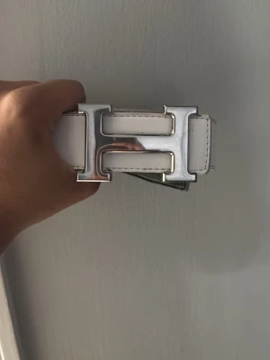 Vitt bälte med H-spänne - Snyggt vitt bälte med ett stort, blankt H-format spänne i silverfärg. Bältet har en stilren och modern look, perfekt för att lyfta vilken outfit som helst. har detaljerade sömmar längs kanterna. Endast Bälte+hole