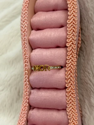 Guldfärgad Dior ring med stenar - Snygg ring från Dior i guldfärg med bokstäverna 'DIOR' framtill. Ringen har små gnistrande stenar på sidorna som ger extra bling. Perfekt accessoar för att lyfta din stil och visa din kärlek för designer.