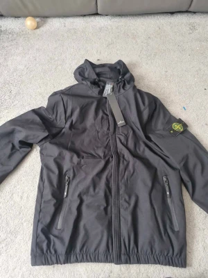 Svart vindjacka från Stone Island - Snygg svart vindjacka från Stone Island med huva och dragkedja framtill. Jackan har två sidofickor med dragkedja och den klassiska Stone Island-loggan på vänster ärm. Materialet är lätt och vindtätt, perfekt för blåsiga dagar.