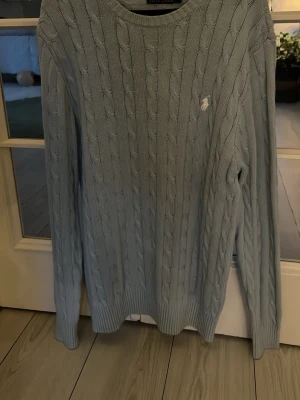 Ljusblå kabelstickad tröja Polo Ralph Lauren - Ljusblå kabelstickad tröja från Polo Ralph Lauren med rund halsringning och klassisk vit broderad logga på bröstet. Tröjan har långa ärmar och ribbade muddar vid ärmslut och nederkant. Perfekt för en stilren och avslappnad look.