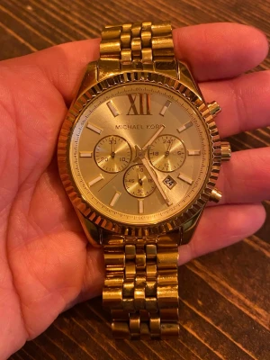 Guldig klocka från Michael Kors - Stilren guldig klocka från Michael Kors med länkarmband och flera små urtavlor. Klassisk design med romerska siffror och datumvisning. Perfekt accessoar för dig som vill sticka ut med en lyxig look.