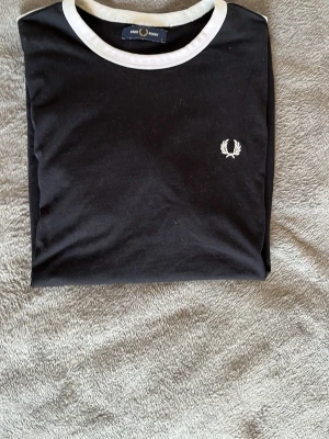 Svart t-shirt med vit krage Fred Perry - Svart t-shirt från Fred Perry med vit krage och vita detaljer vid ärmslut. Klassisk logga broderad på bröstet. T-shirten har en normal passform och är tillverkad i mjuk bomull. Perfekt för en clean och stilren look.