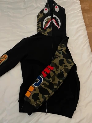 Svart Camo Bape Hoodie - Säljer en svart Bape hoodie med dragkedja och huva. Ena ärmen och huvan har camomönster och färgglada bokstäver, samt ett hajansikte med tänder och ögon på huvan. Tröjan är mycket för kort i längd och ärmar för modellen på bilden som är 177cm lång och väger 65kg för referens.