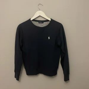 Blå tröja från Polo Ralph Lauren - Klassisk mörk blå tröja från Polo Ralph Lauren med rund halsringning och diskret broderad logga på bröstet. Passar dig runt 140-152cm lång. Långärmad modell i mjukt material, perfekt för en clean och stilren look. Passar dig som gillar enkel men snygg design.