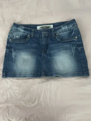 Blå jeanskjol med slitningar - Snygg blå jeanskjol i kort modell med slitna detaljer och klassiska fem fickor. Kjolen har coola detaljer bak. Perfekt för en avslappnad och trendig look. Kjolar har ingen knapp men är fixat med en metal grej som syns på sista bilden. Jag kan prata om priset. 