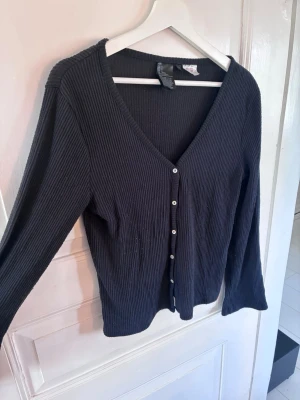 Svart/grå ribbad kofta från H&M - Stilren svart/grå ribbad kofta från H&M med v-ringning och långa ärmar. Koftan har knäppning framtill med vita knappar och är tillverkad i ett mjukt, stretchigt material som sitter snyggt på kroppen. Perfekt att slänga över en topp eller klänning.