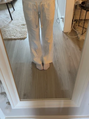 Vita linnebyxor med dragsko - Snygga vita linnebyxor med avslappnad passform och bred resår i midjan. Byxorna har dragsko och stora fickor framtill. Perfekta för en chill och stilren look. Materialet är luftigt linne som känns fräscht och bekvämt. 