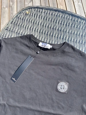 Svart Stone Island t-shirt - Snygg svart t-shirt från Stone Island med klassisk logopatch på bröstet. Tillverkad i mjuk bomull och har rund hals samt korta ärmar. Perfekt för dig som gillar stilrena och ikoniska plagg.