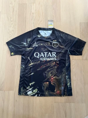 Svart PSG matchtröja med marmorprint - Säljer en svart PSG matchtröja från Jordan med unikt marmorliknande print i guld och grått. Tröjan har korta ärmar, rund hals och PSG-logga samt Jordan-logga på bröstet. Tillverkad i lätt och ventilerande funktionsmaterial. Perfekt för fotboll eller streetwear.