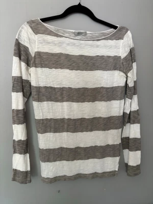Randig Hunkydory topp med vingar - Säljer en långärmad Hunkydory topp med breda grå och vita ränder. Toppen har en rund halsringning och är dekorerad med broderade vita vingar på ryggen. Tillverkad i ett mjukt och tunt bomullsmaterial som känns skönt mot huden.